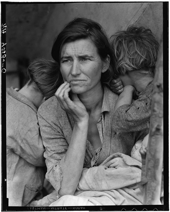 Black-and-white photograph of a worried woman in worn clothes, one hand at her chin, with two children leaning on her shoulders and their faces turned away, and a baby in her lap.